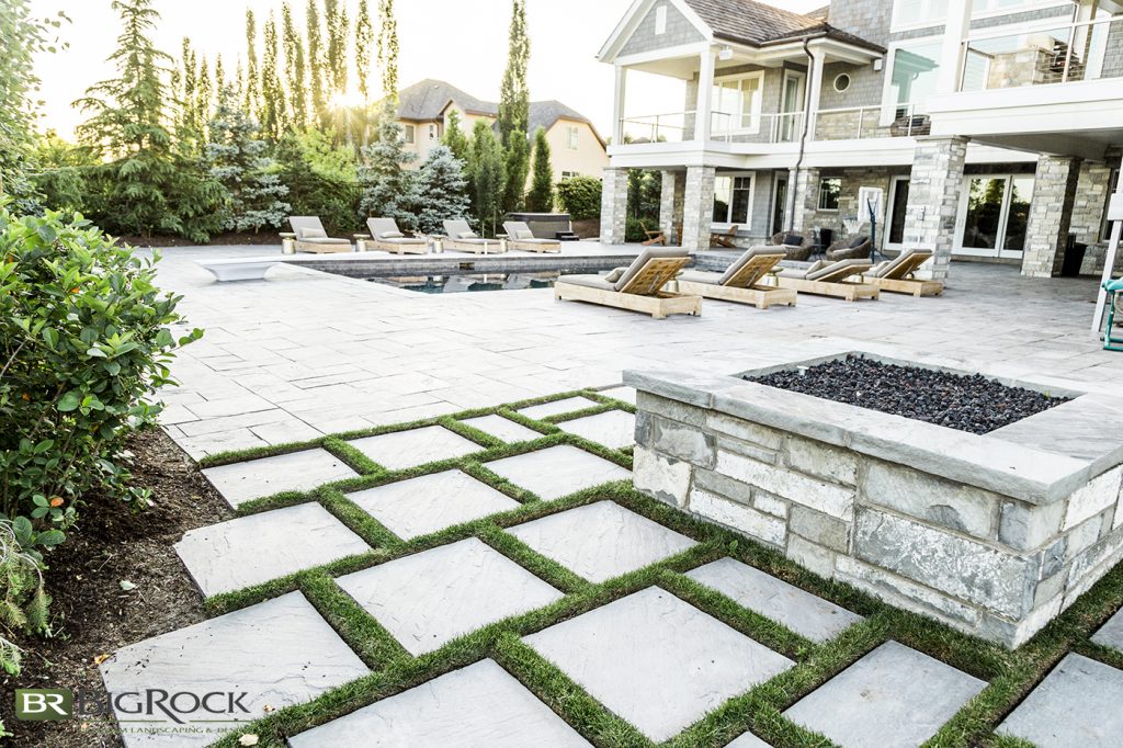 A gray stone is used on the patio decking, the firepit, and on the house itself, which lends a unifying element to the space, while the uneven stonework on the firepit and house contrasts with the smooth surface of the pool decking