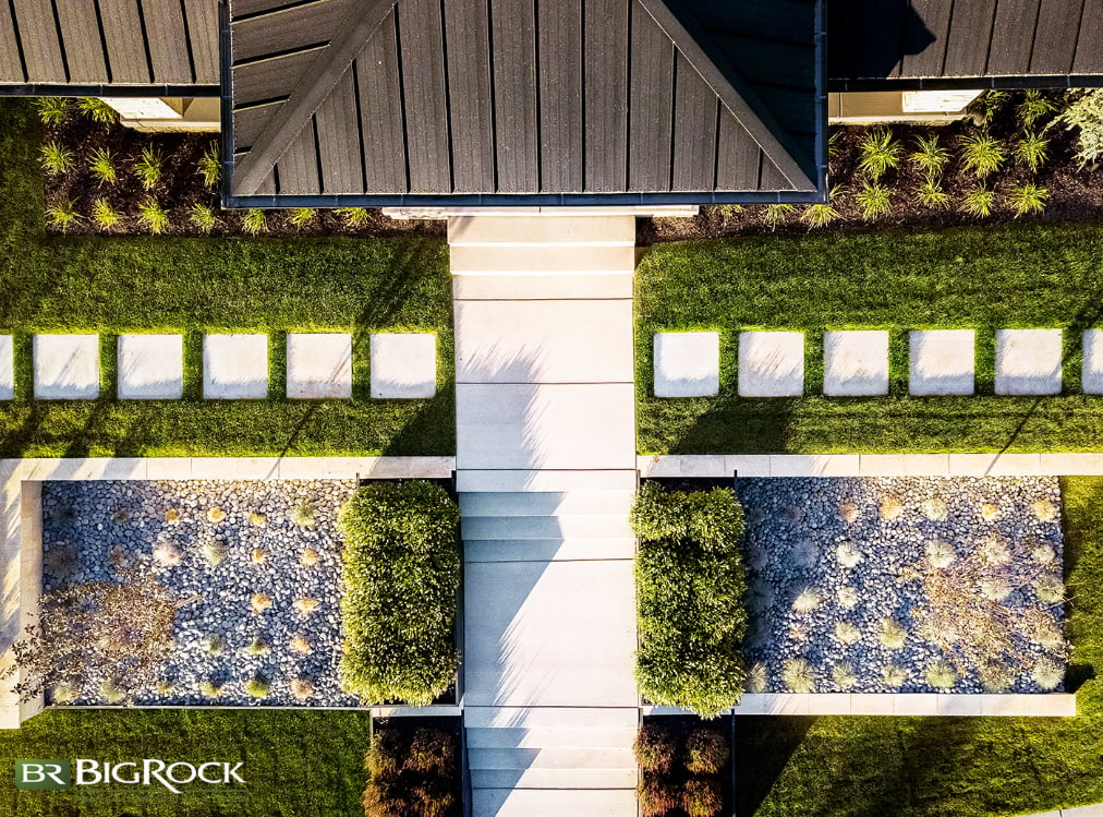 Every element in this sloped front yard exemplifies modern landscaping. The clean lines and the bright white of the pavers, the river rock in the flower beds, trees planted in symmetrical places, boxwoods lining the front walk, and blue fescue as the only plant in the beds.