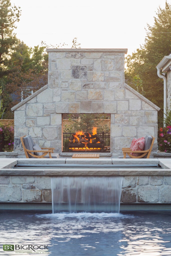 This hardscape design enhances the pool space in three ways: with a fireplace, hot tub, and waterfall. The firepit near the hot tub makes this space usable year-round, making it easy to enjoy a winter dip.